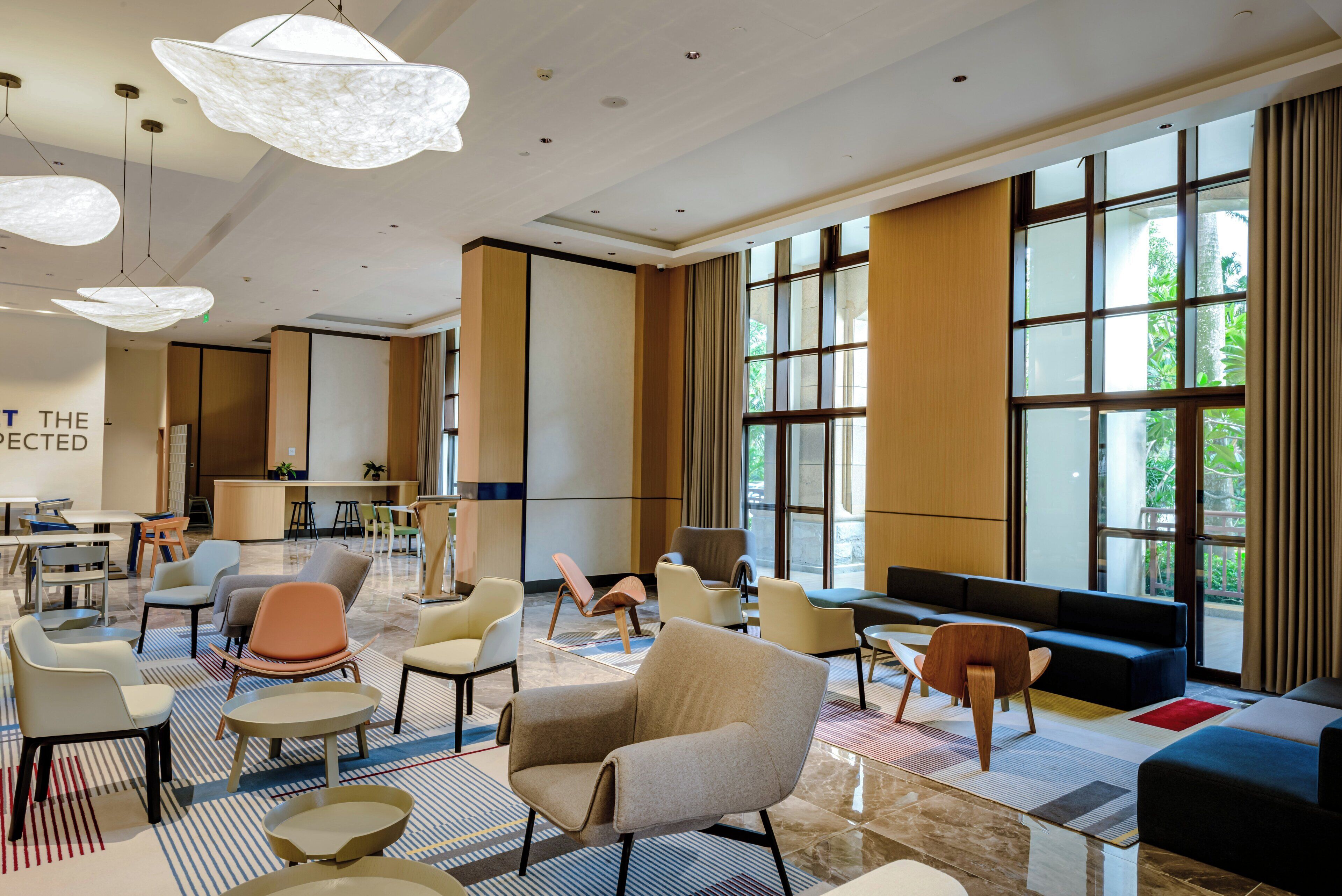 A modern hotel lobby with a mix of seating areas, including sofas and chairs, a reception desk, and large windows offering views of the outdoors.