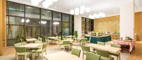 A modern dining area with multiple tables and chairs, large windows, and a kitchen area in the background.