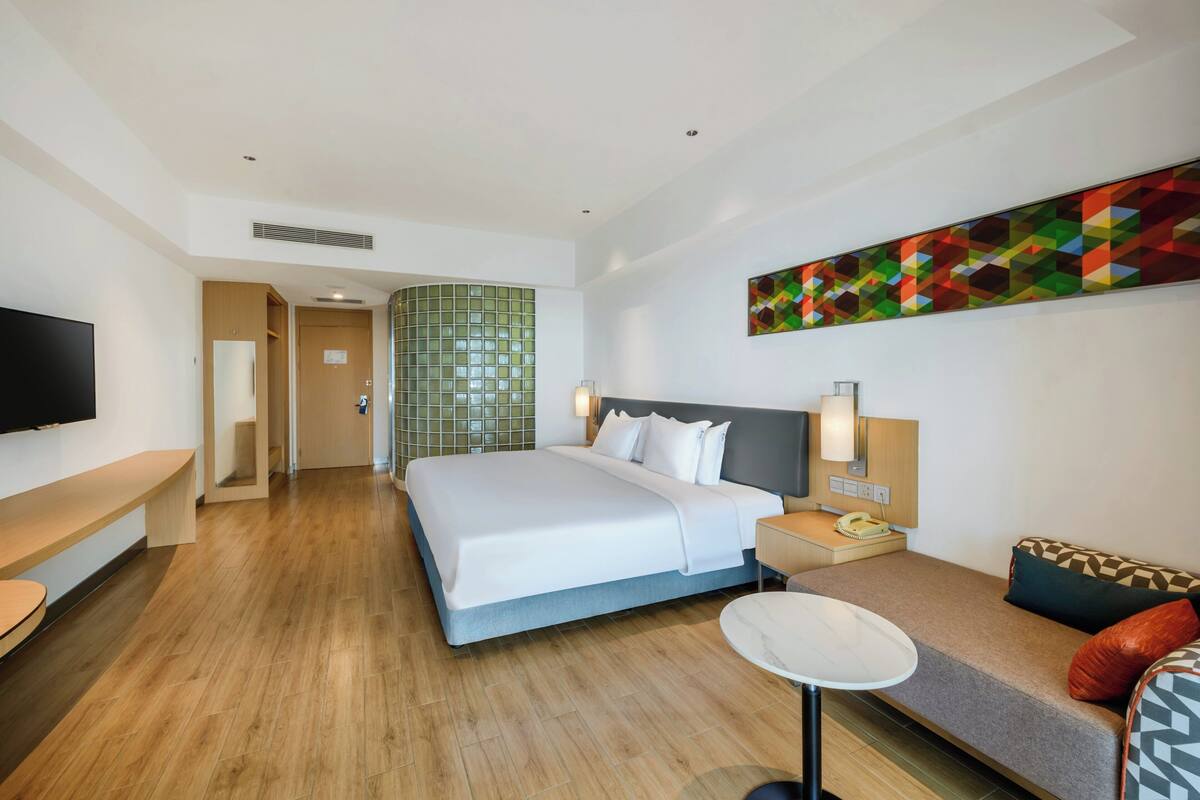 Habitación de hotel con una cama grande, una mesita de noche con lámpara, un pequeño sofá, un televisor y un cuadro abstracto colorido en la pared.