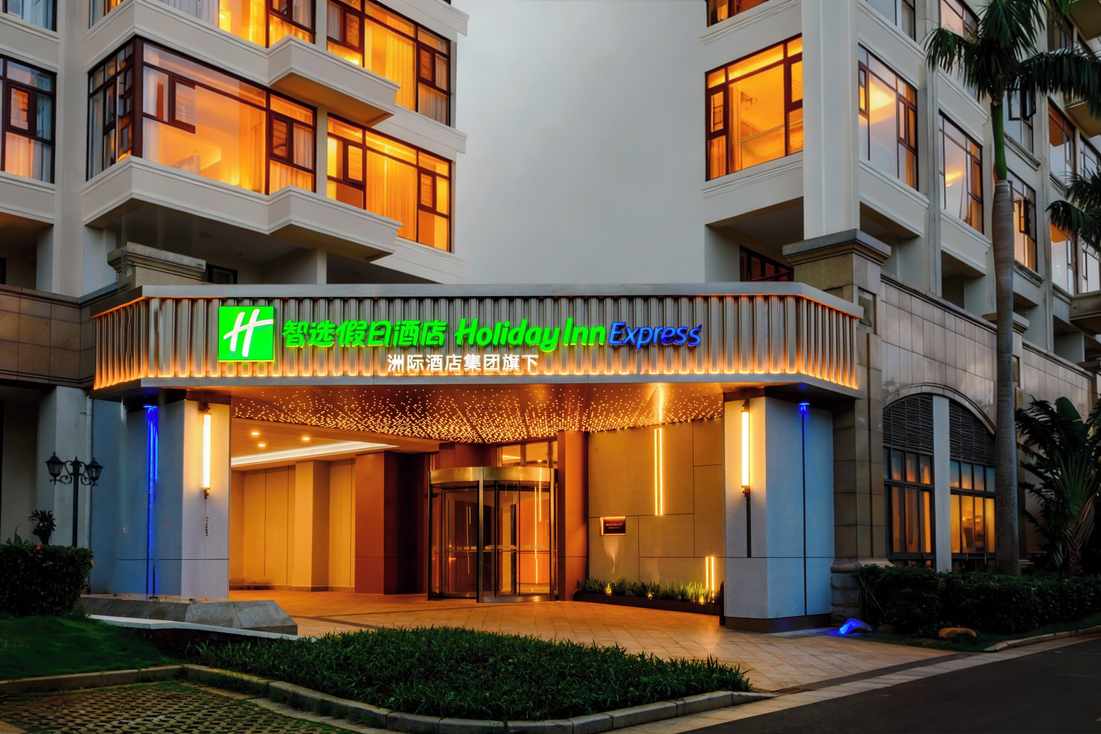 A hotel entrance with illuminated signage, a gated entrance, and a landscaped area with a pathway.