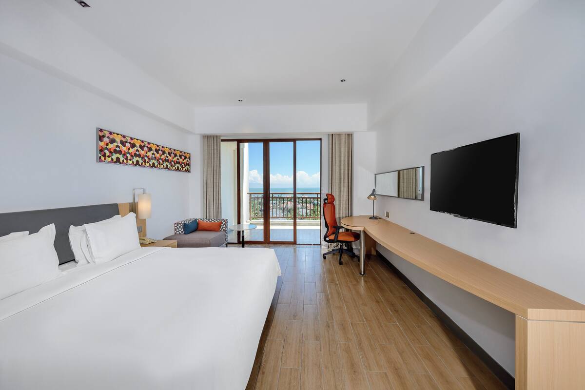 Habitación de hotel con una cama grande, un escritorio, una silla, un televisor y un balcón con vistas.