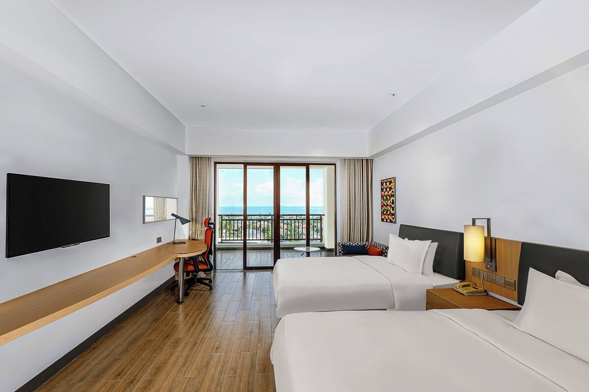 Habitación de hotel con televisión, escritorio, dos camas, balcón con vistas y mesita de noche con lámpara.