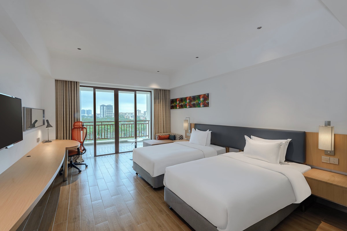 Een hotelkamer met twee bedden, een bureau, een televisie en een balkon met uitzicht op de stad.