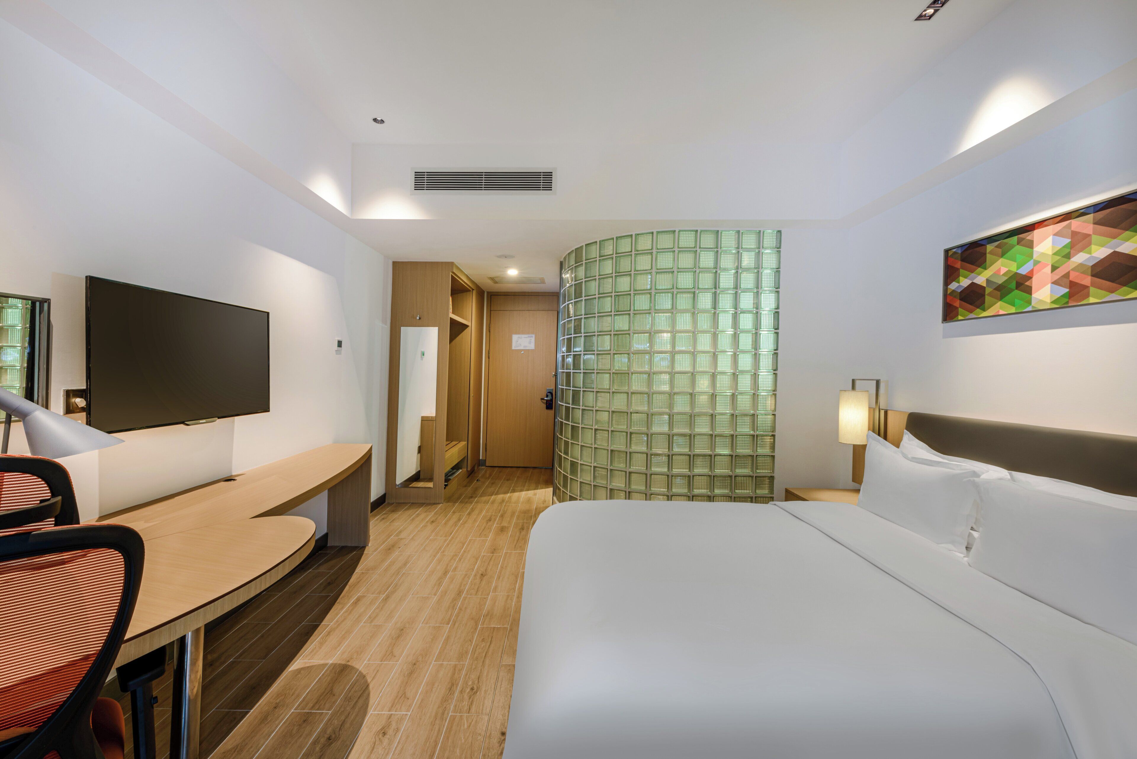A hotel room with a bed, a desk, a chair, a TV, a green patterned wall, and a painting.