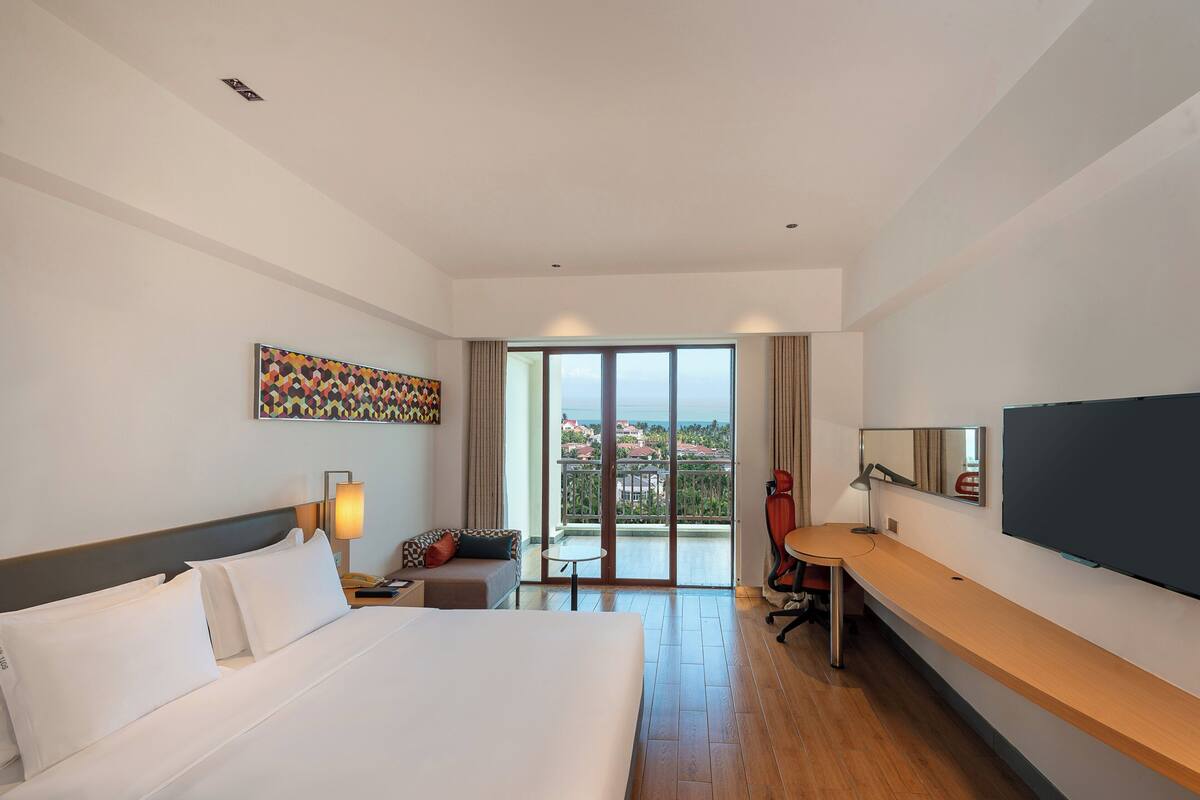 A hotel room with a bed, a desk, a chair, a TV, a painting, and a balcony with a view.