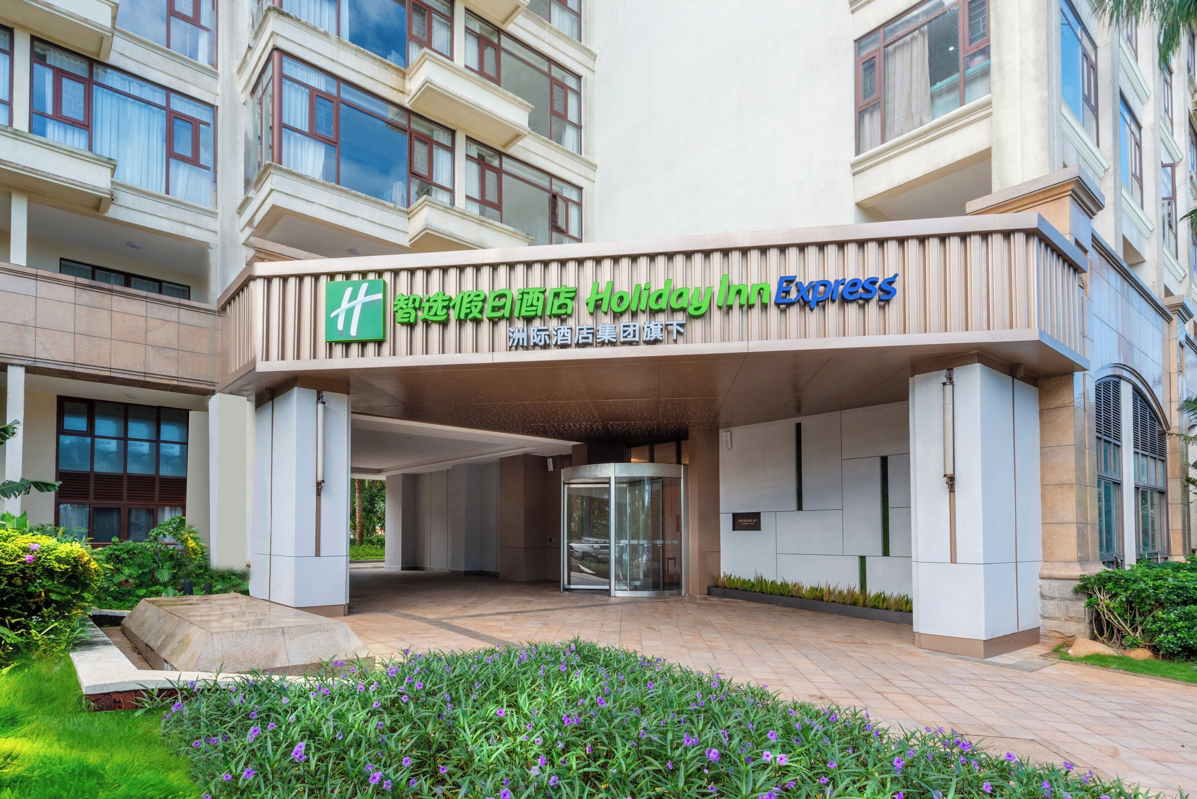 A Holiday Inn Express hotel entrance with a modern design, a revolving door, and a landscaped area with flowers.