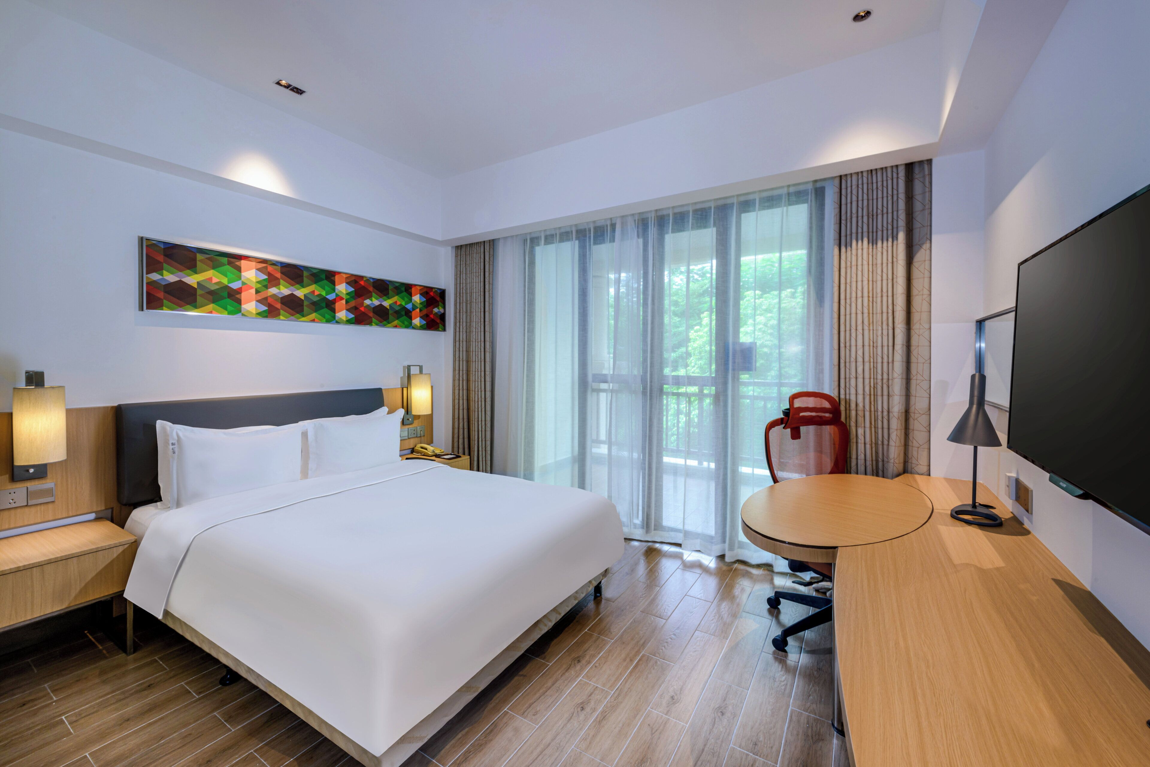Quarto de hotel moderno com cama grande, escrivaninha, cadeira, televisão e janela com cortinas.