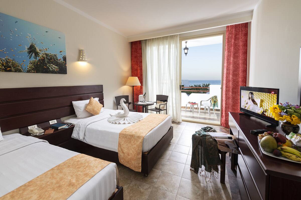 A hotel room with two beds, a TV, a desk with a lamp, and a view of the ocean.