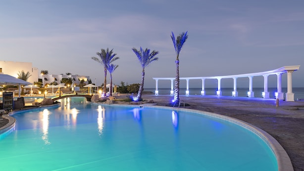 A swimming pool with palm trees and a covered area, surrounded by buildings and a view of the sea. - Three Corners Equinox Beach Resort (Marsa Alam)