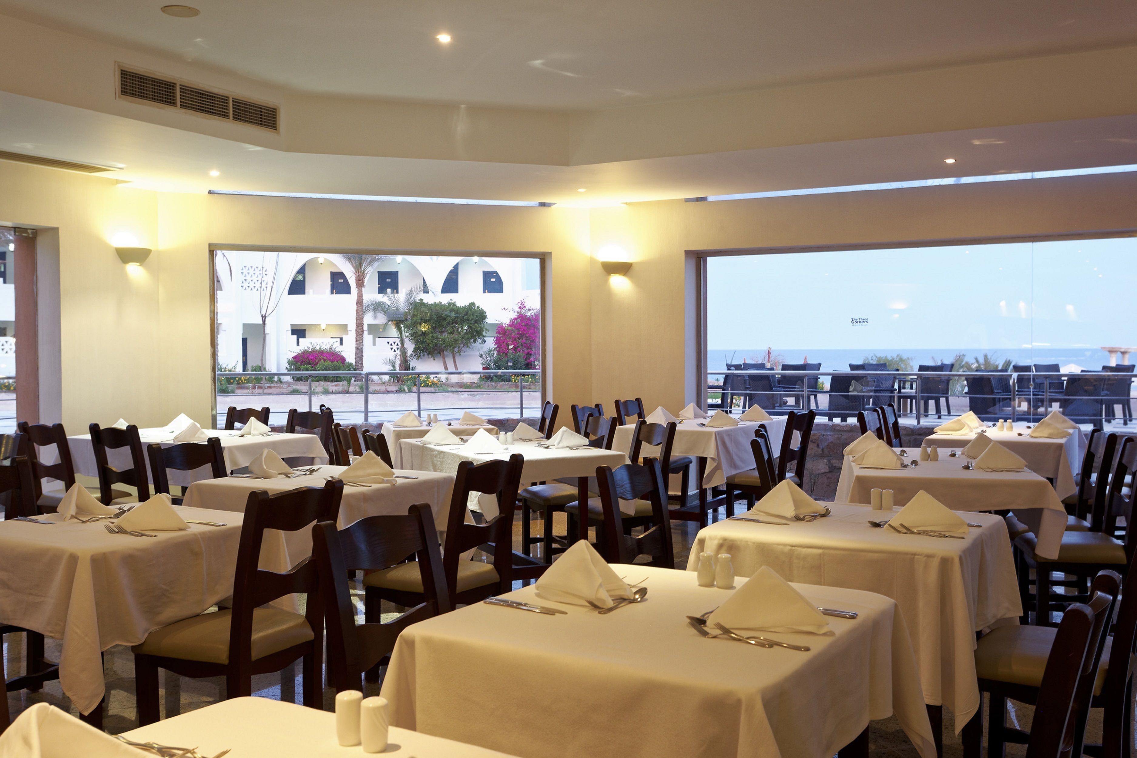 Un restaurante con varias mesas dispuestas para comer, amplios ventanales que ofrecen vistas al mar y a edificios, y una decoración interior moderna.