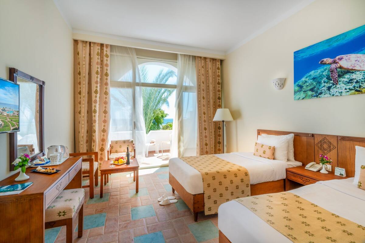 A hotel room with two beds, a desk with a chair, a small table with a vase of flowers, a lamp, and a painting of a sea turtle on the wall.