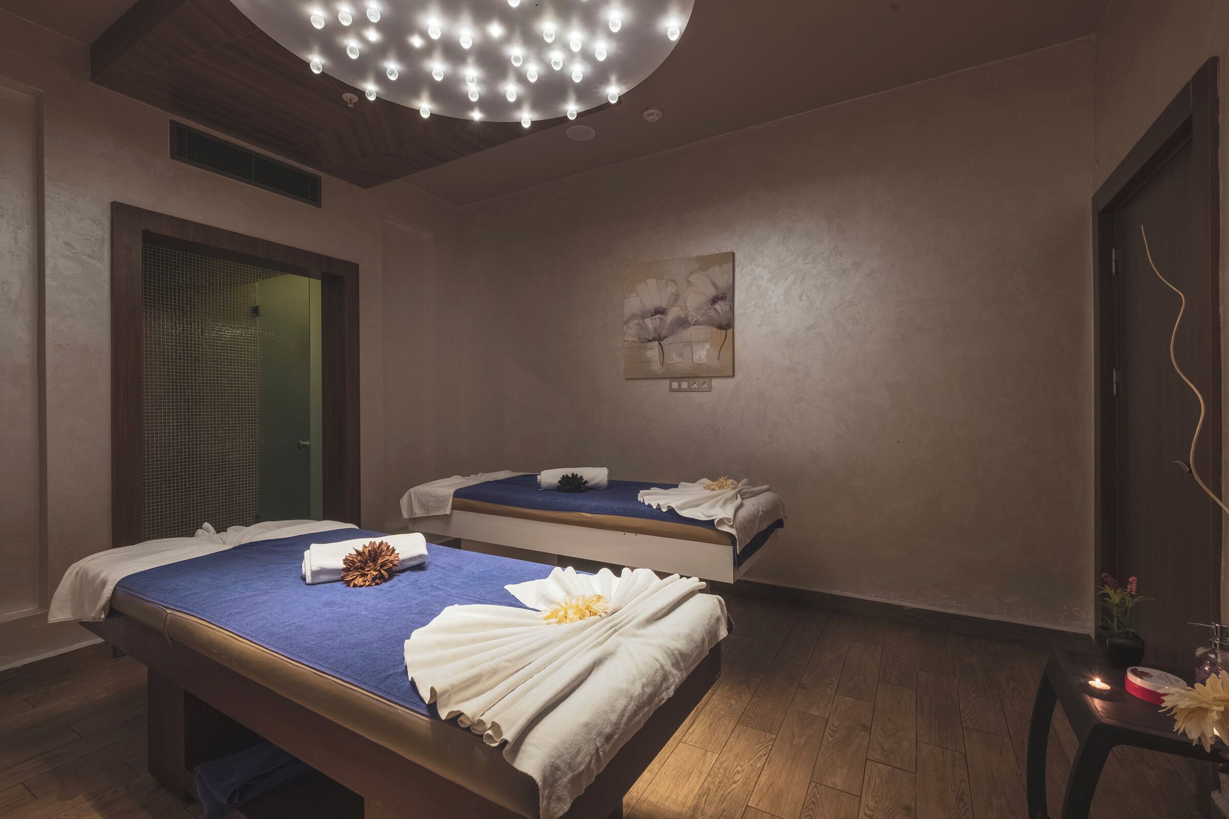 A spa room with two beds, each with a blue cover and white towels. There is a ceiling light fixture, a painting on the wall, and a small table with a candle and flowers.
