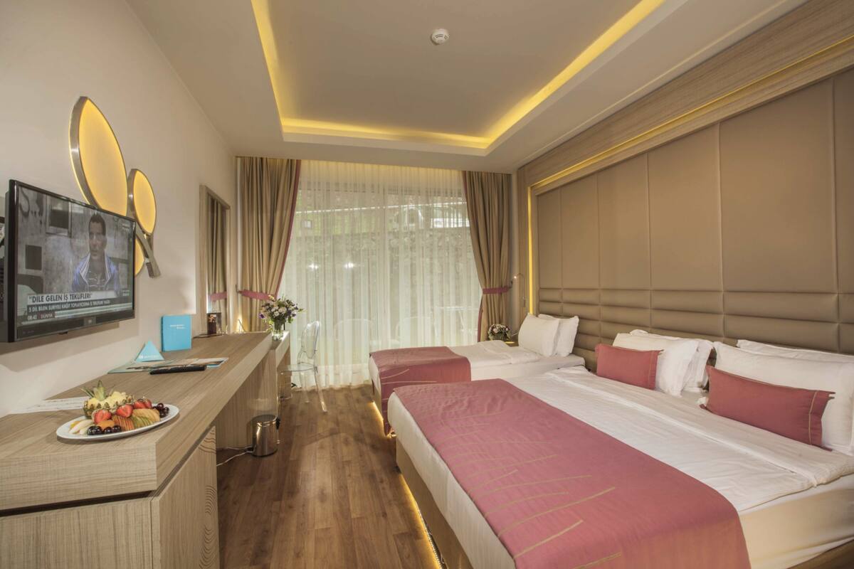 Une chambre d’hôtel avec deux lits, un bureau sur lequel se trouve une assiette de fruits, une télévision et une grande fenêtre avec des rideaux.
