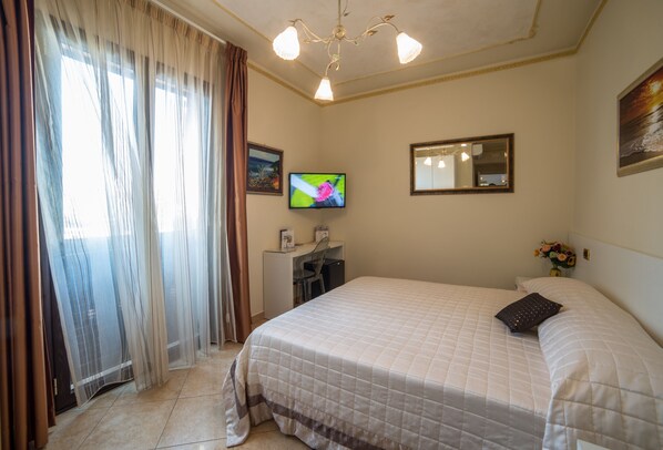 Habitación de hotel con cama, escritorio, televisor, espejo y ventana con cortinas.
