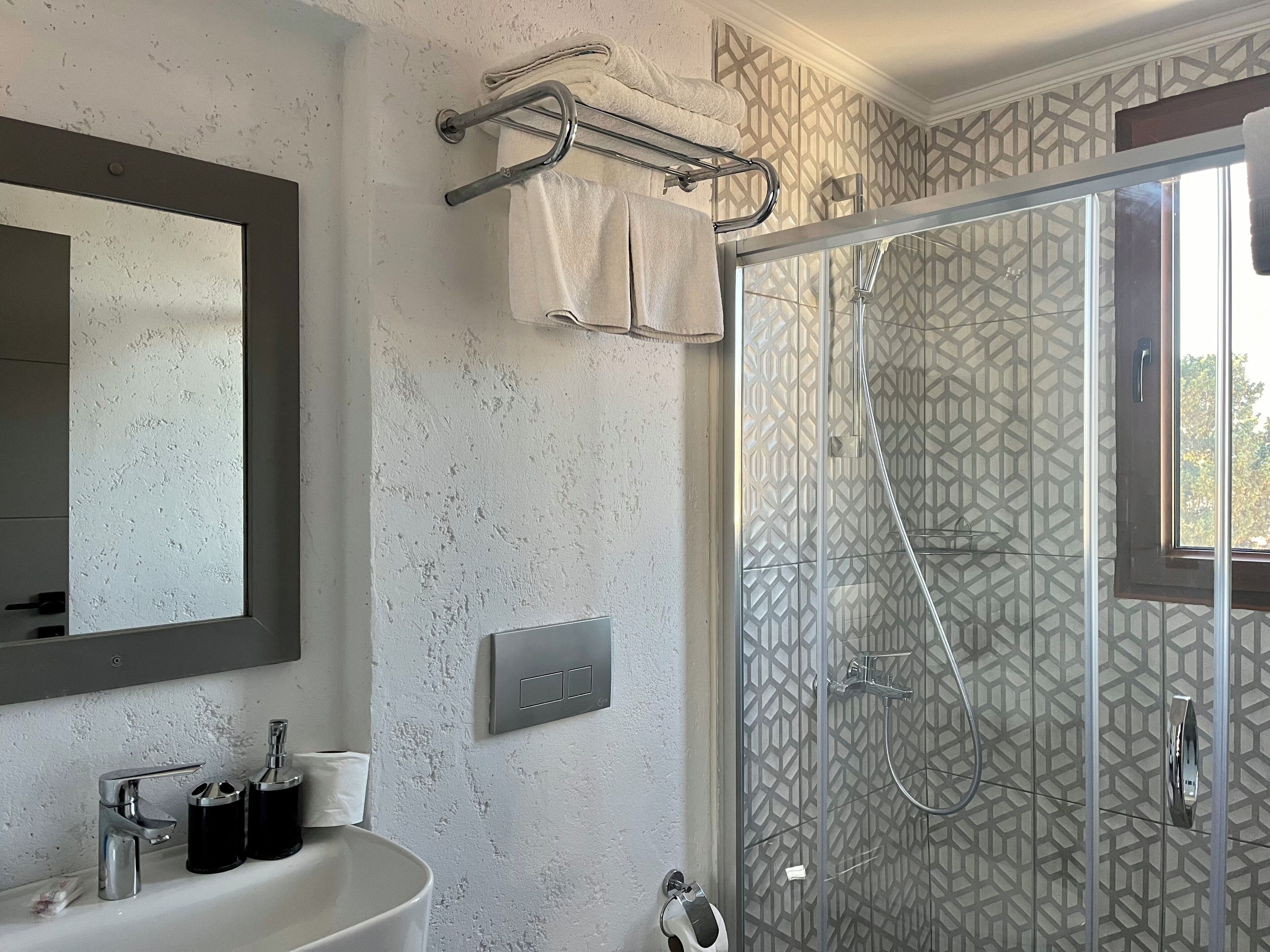 A bathroom with a large mirror, a sink with two faucets, a towel rack, and a shower with a geometric-patterned glass door.