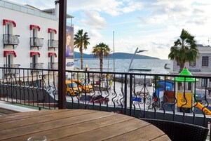 Un balcón con vista al frente marítimo, que incluye una mesa y sillas de madera, barandilla metálica y un edificio con balcones y un mural.