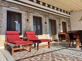 Un espacio de estar al aire libre cubierto con dos sillones de madera y una mesa, frente a una pared de piedra con ventanas amplias.