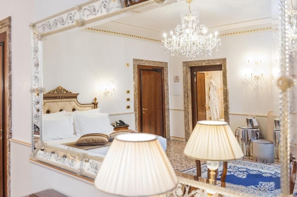 A luxurious bedroom with a large ornate mirror, a chandelier, and a bed with white linens.