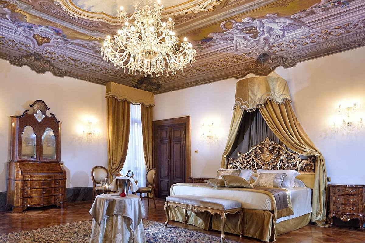 Una habitación amplia con un techo ornamentado, un candelabro, una cama grande con dosel, un aparador de madera y una mesa con mantel.