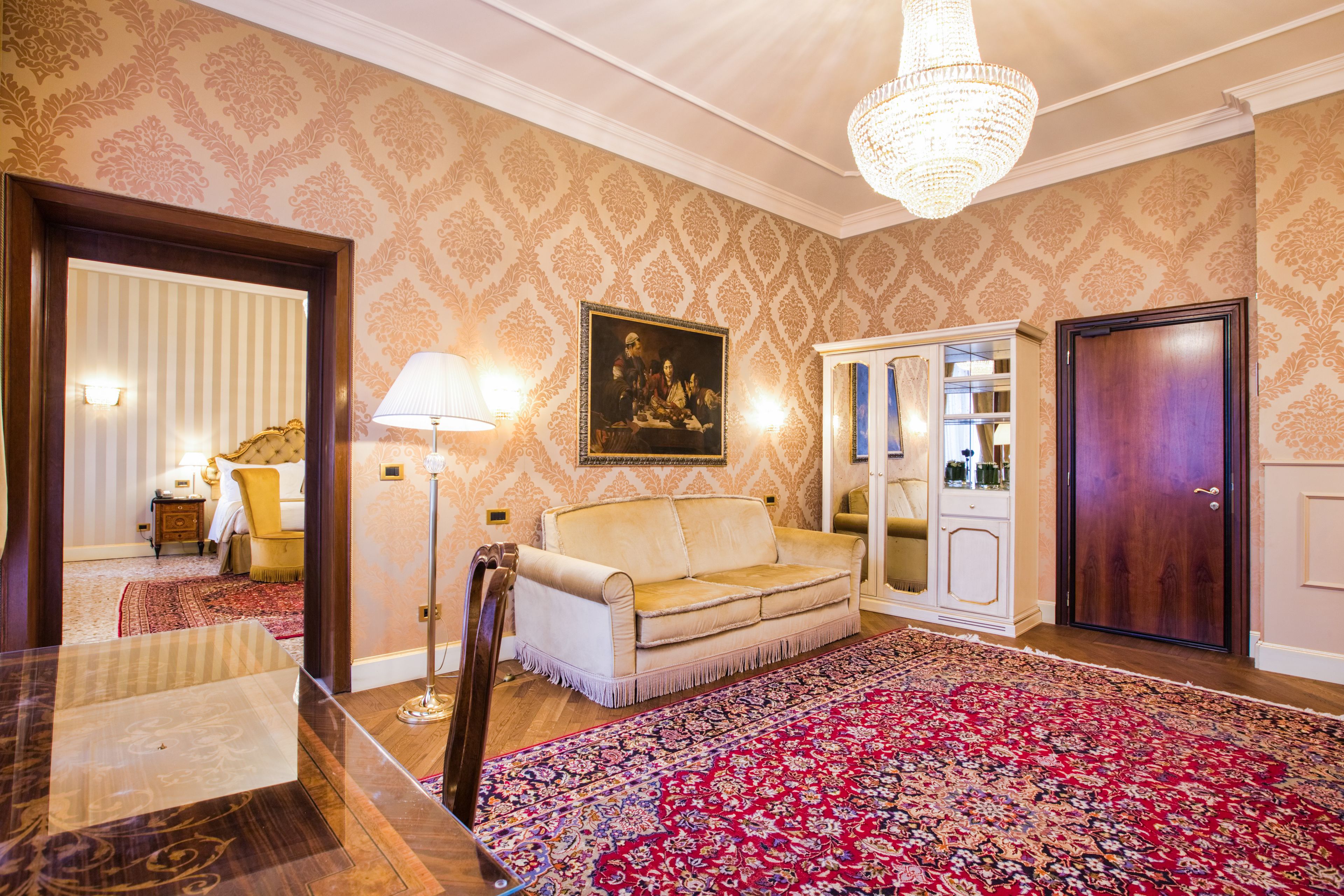 A room with a patterned wallpaper, a chandelier, a sofa, a cabinet, and a large rug.
