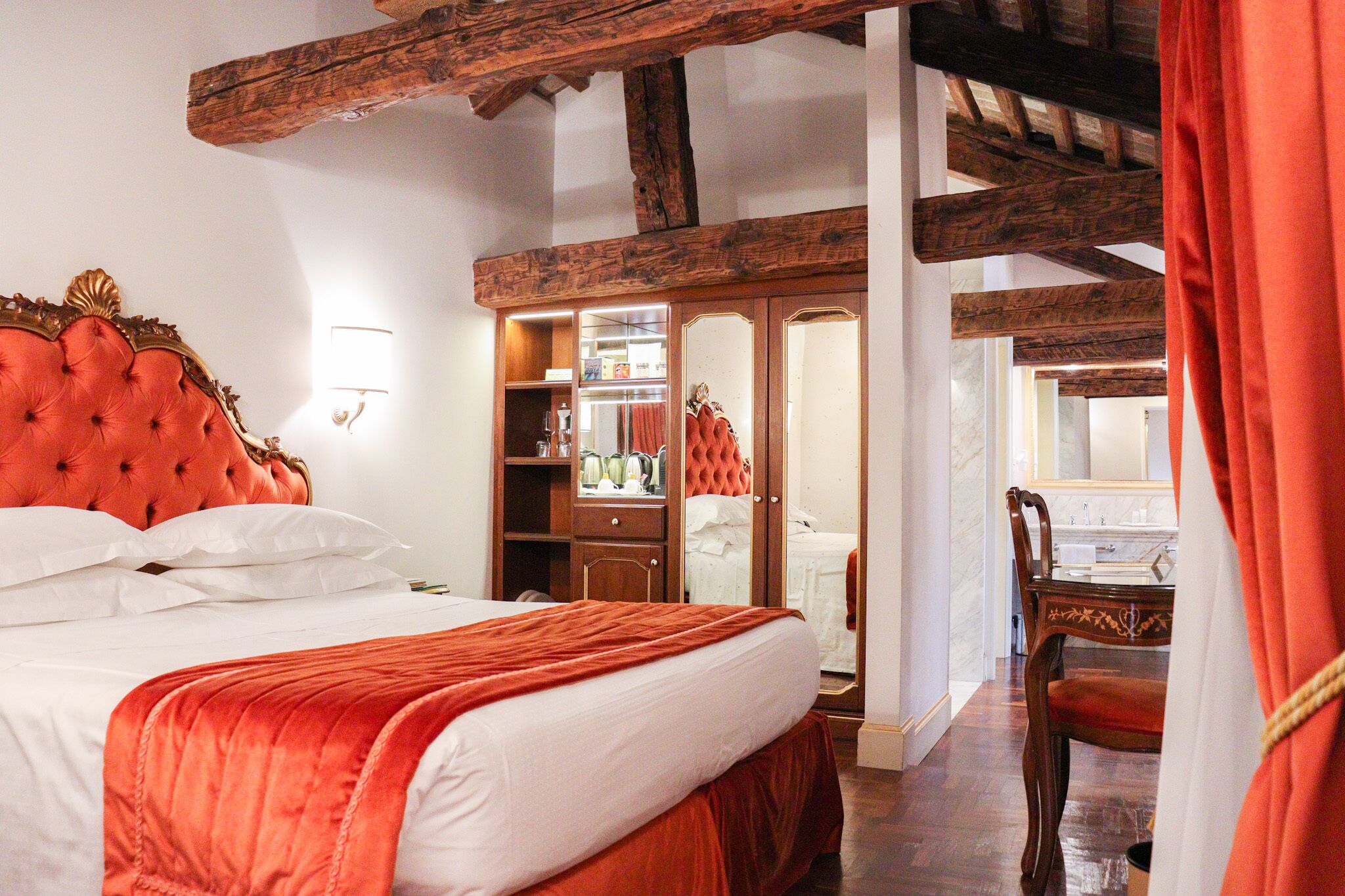 Un dormitorio con una cama grande, vigas de madera en el techo, un armario y un baño visible a través de una puerta abierta.