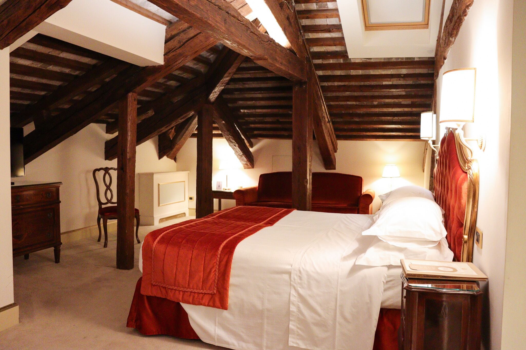 Une chambre avec un plafond en bois, un lit recouvert d’une couverture rouge, une chaise, une table de chevet et une lampe.