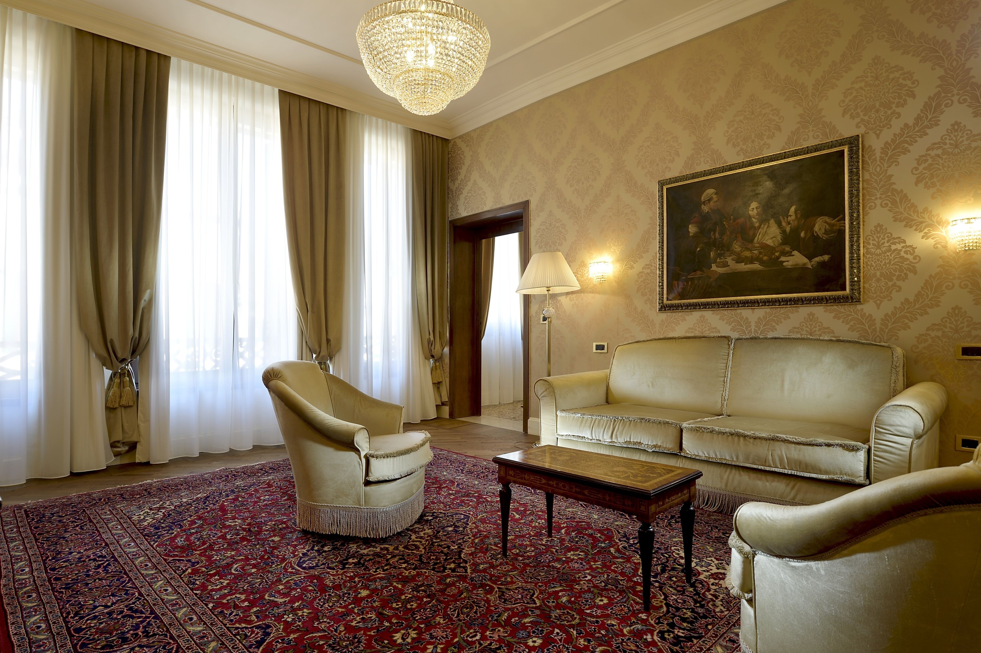 A room with a large chandelier, a sofa, a chair, a patterned rug, and a painting on the wall.