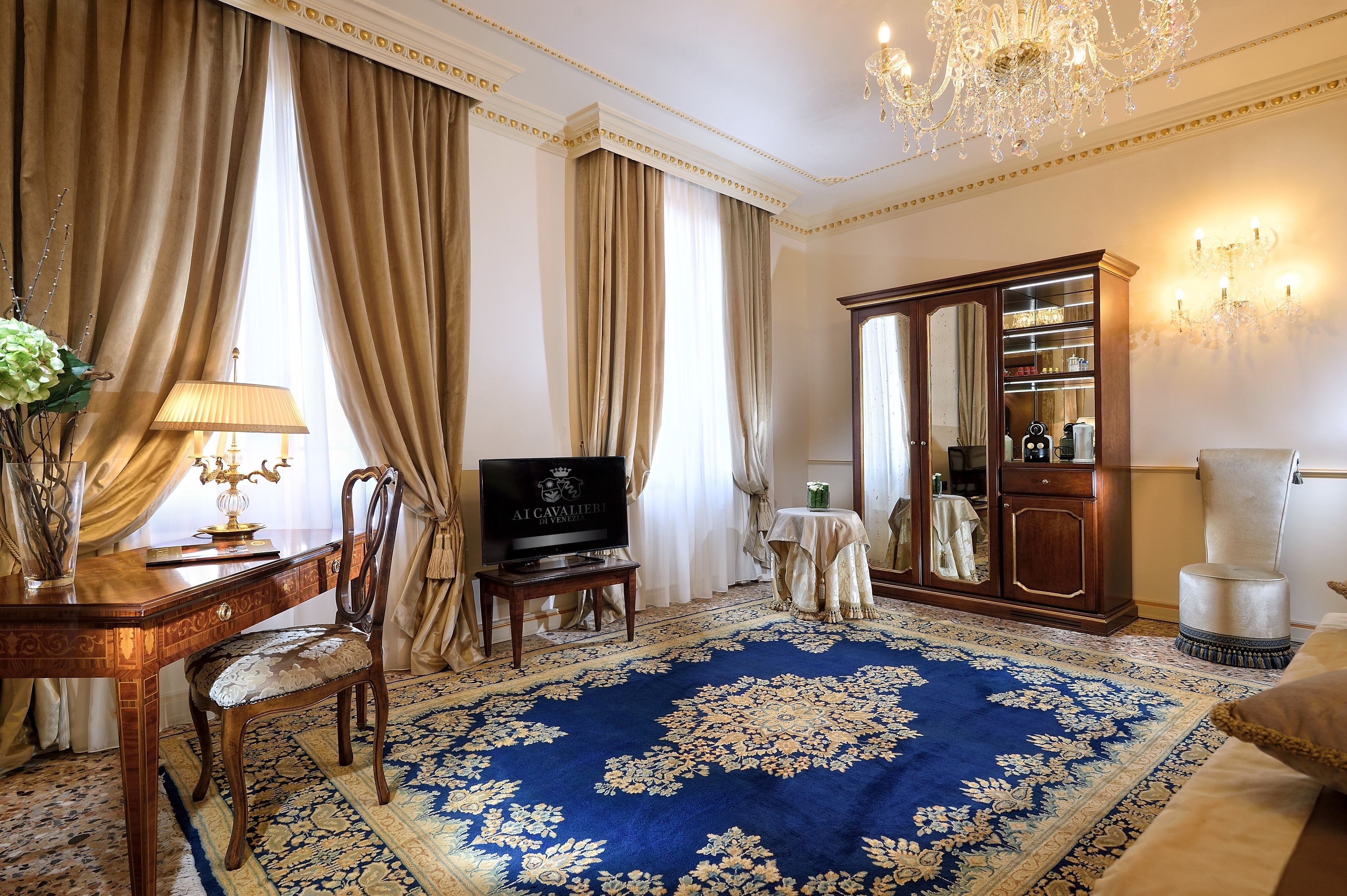 A room with a large blue patterned carpet, a wooden desk with a television, a chair, a cabinet with glass doors, and a chandelier.