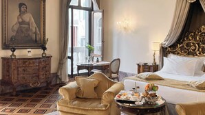 Habitación de hotel con cama, sofá, mesa de comedor con desayuno, cómoda de madera y un amplio ventanal con cortinas.