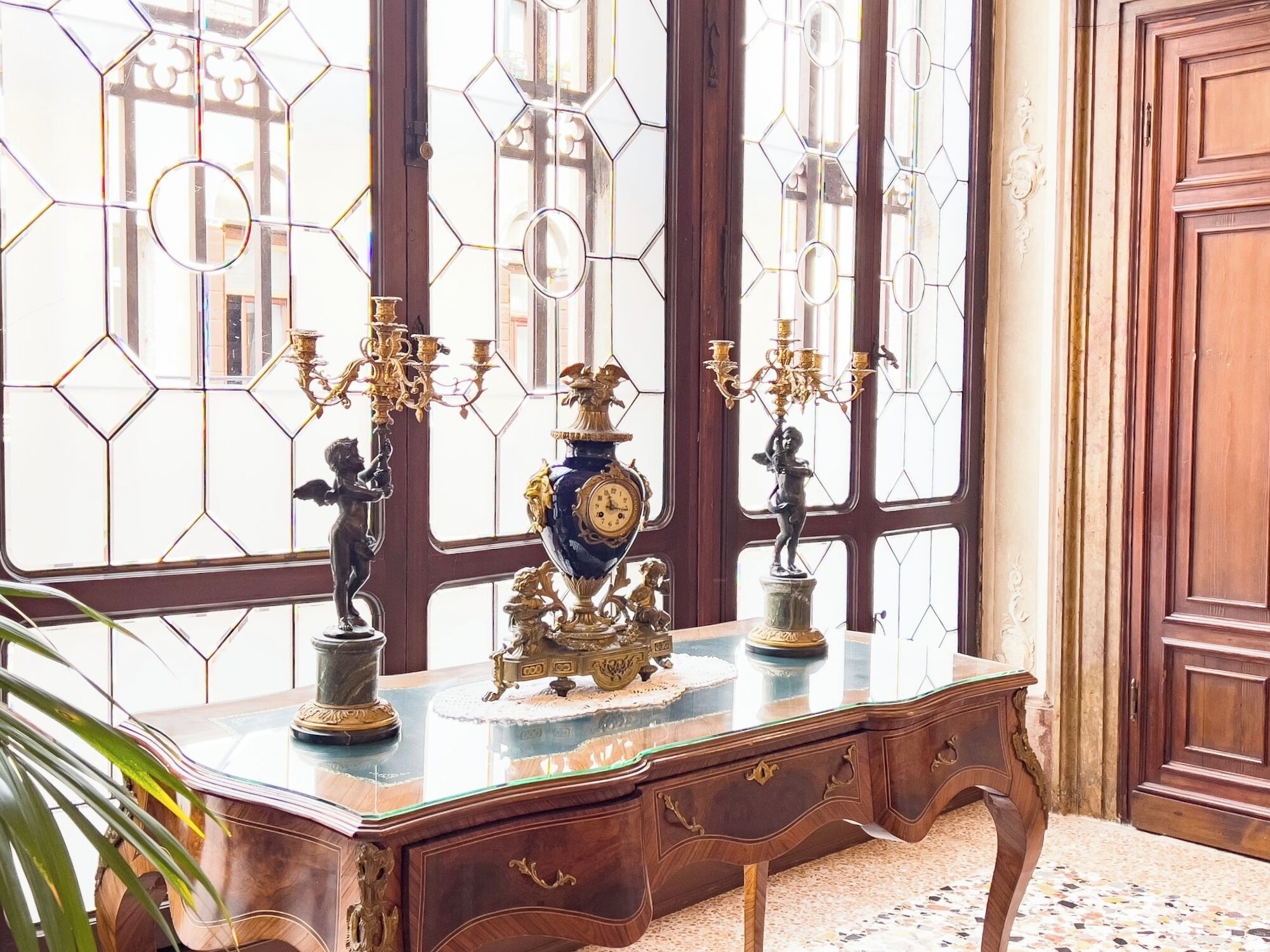 A wooden desk with a glass top, surrounded by ornate wall sconces and a large vase.