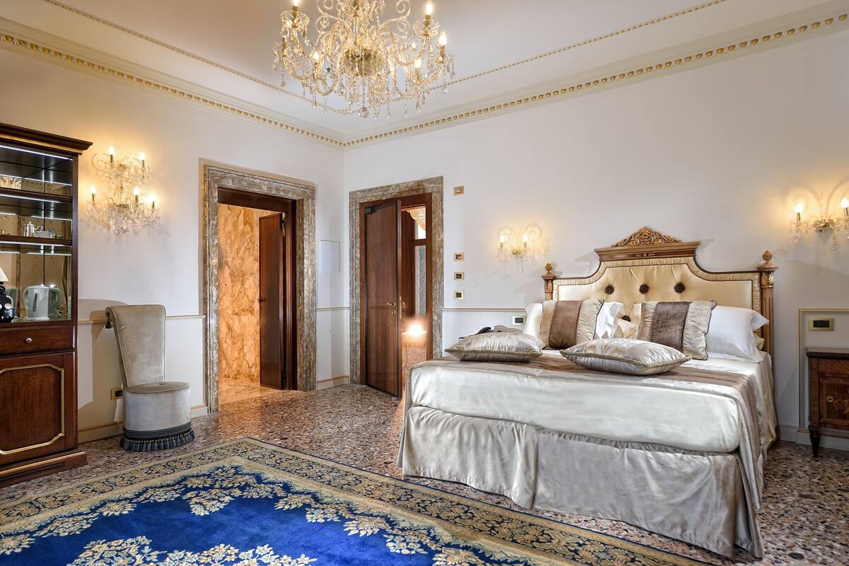 Un dormitorio lujoso con una cama grande, una lámpara de araña ornamentada y una alfombra azul con motivos.
