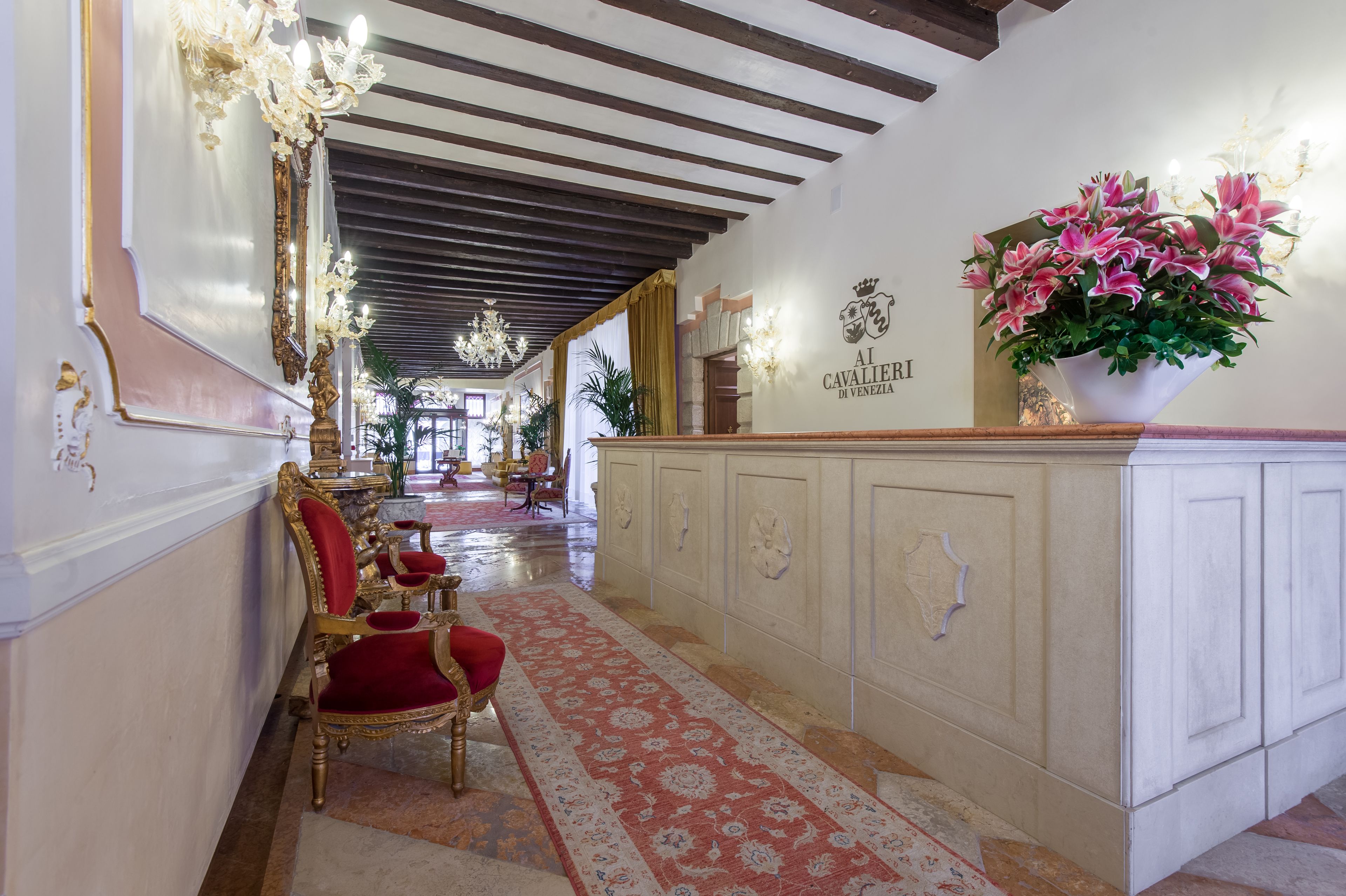 A grand hallway with ornate chandeliers, a red velvet chair, and a white cabinet with a floral arrangement.