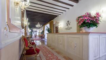 A grand hallway with ornate chandeliers, a red velvet chair, and a white cabinet with a floral arrangement.
