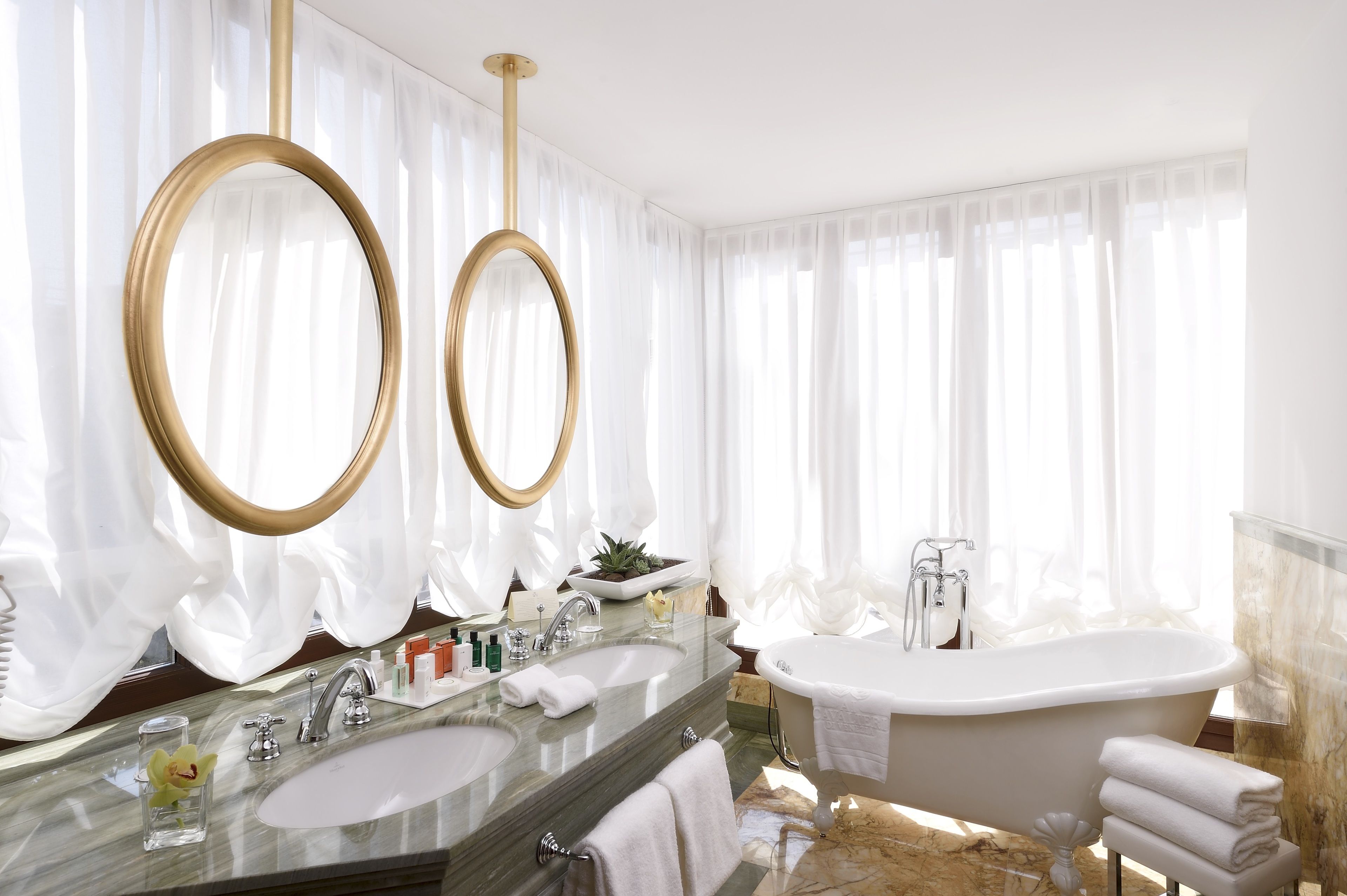A luxurious bathroom with two large mirrors, a freestanding bathtub, a marble sink, and a large window with sheer curtains.