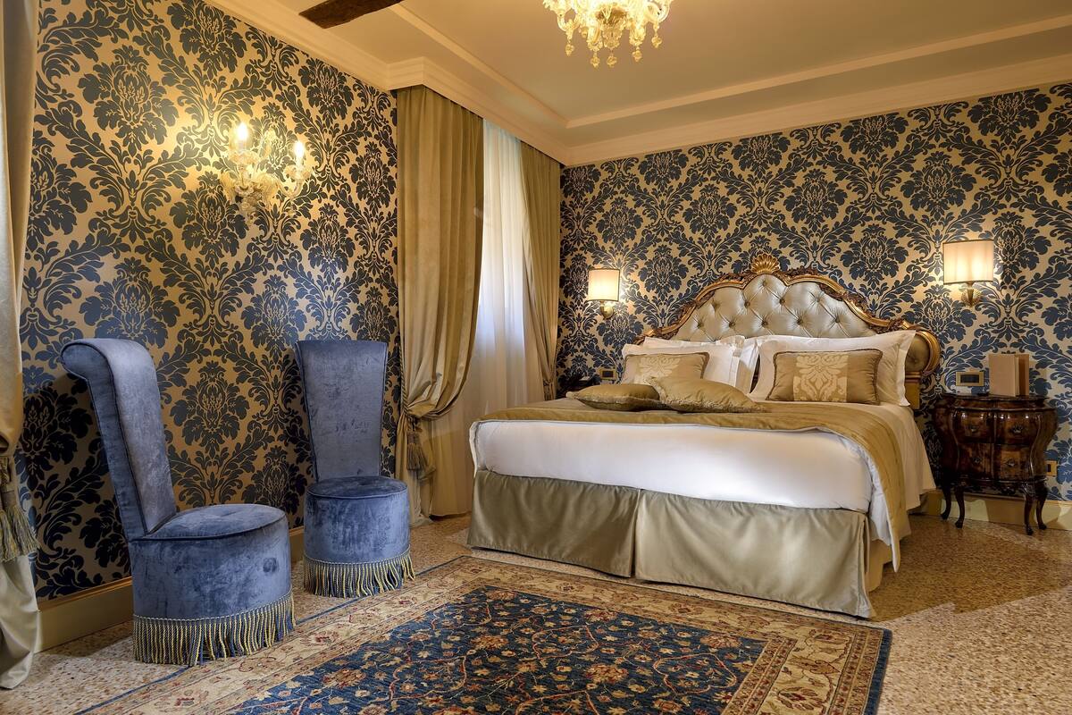 Un dormitorio con una cama grande, dos sillones, una araña y papel tapiz estampado.