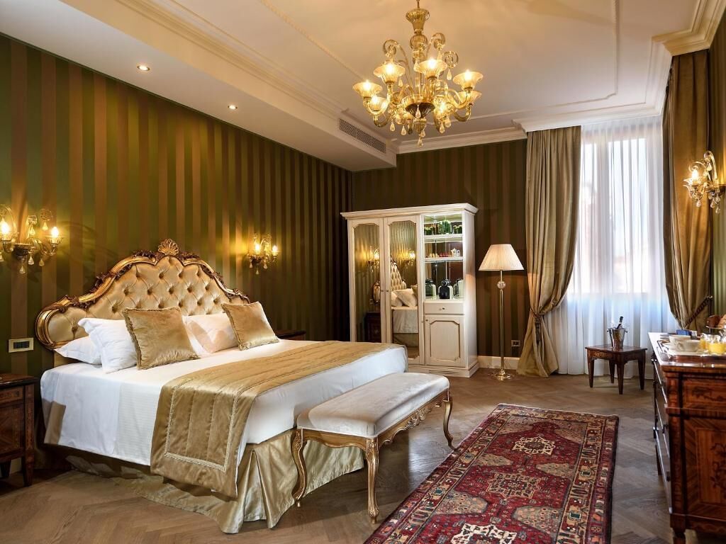 A luxurious bedroom with a large bed, ornate headboard, a chandelier, a cabinet with glass doors, and a red patterned rug.