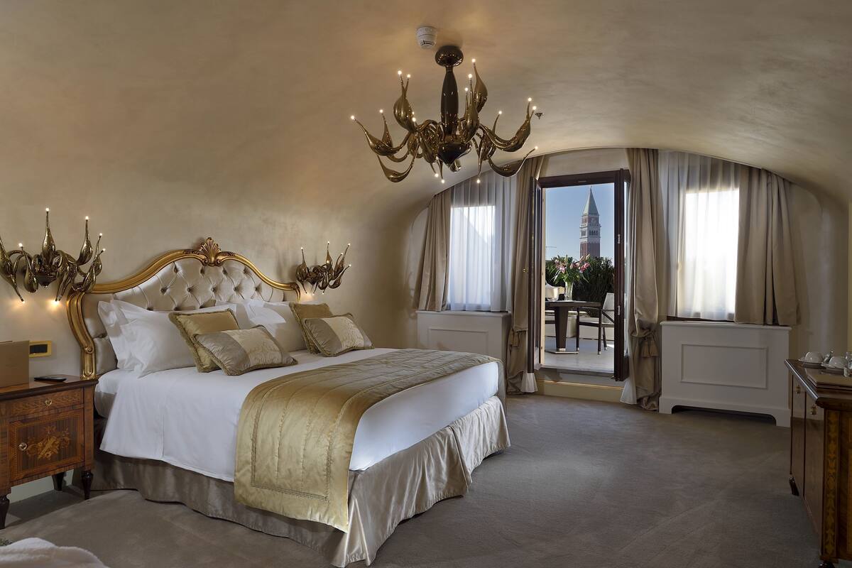 Un dormitorio lujoso con una cama grande, una lámpara de araña ornamentada y vistas a una torre a través de una ventana.