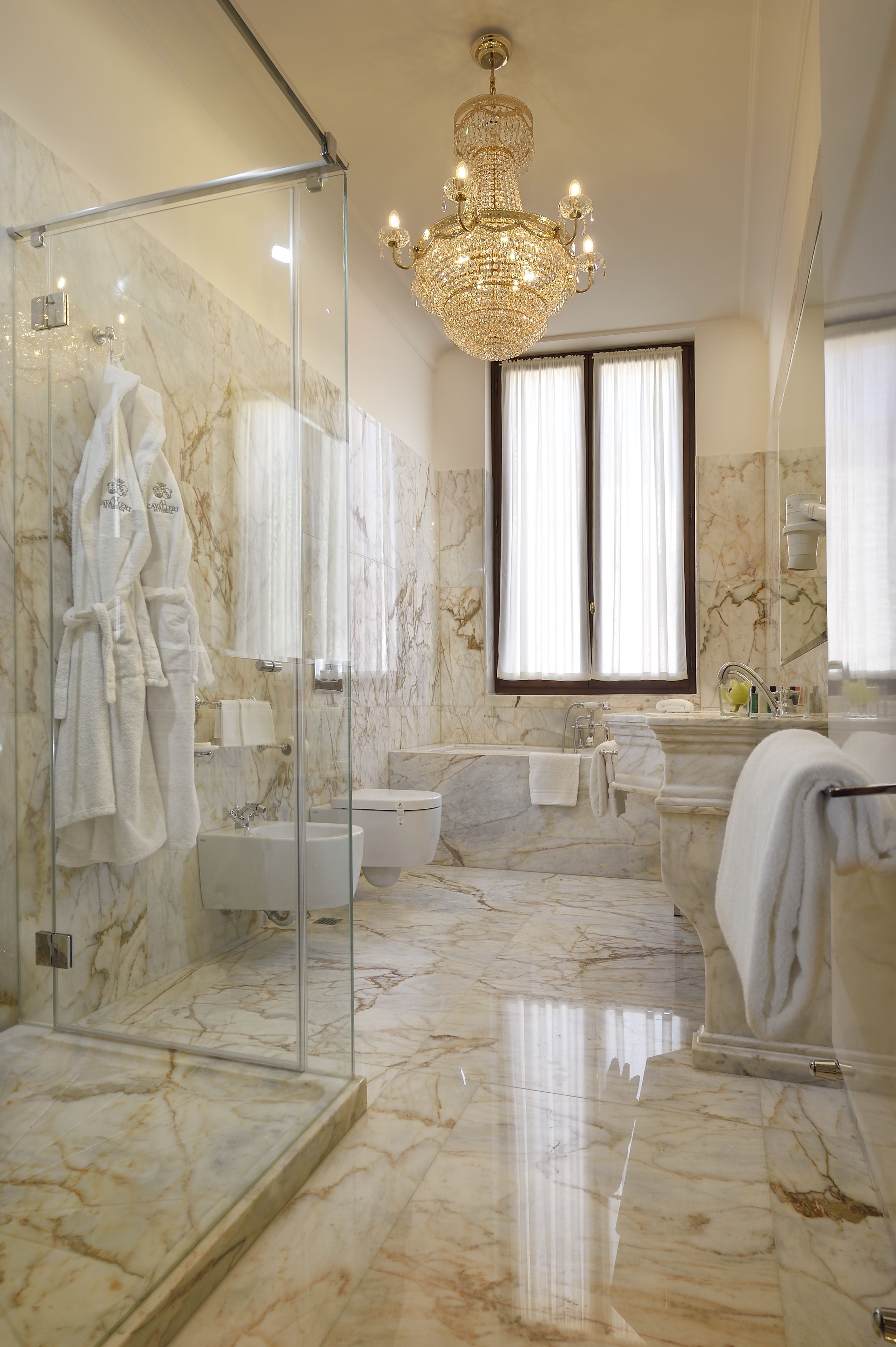 A luxurious bathroom with a glass-enclosed shower, a marble floor, a large chandelier, and a bathtub.