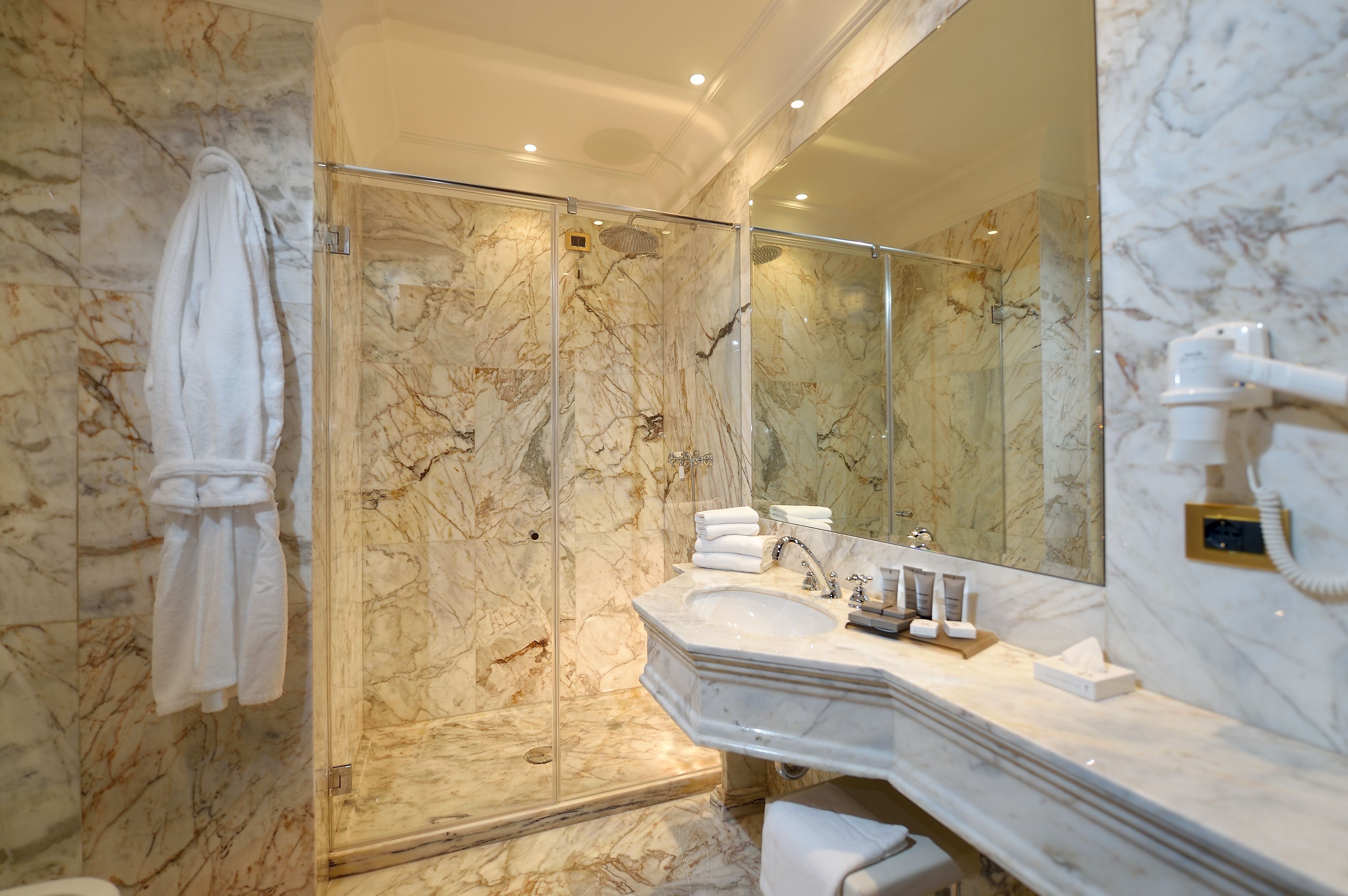 A marble bathroom with a glass-enclosed shower, a large mirror, and a sink with toiletries.