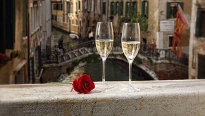 Un balcón con dos copas de vino blanco y una rosa roja, con vista a un canal y edificios al fondo.