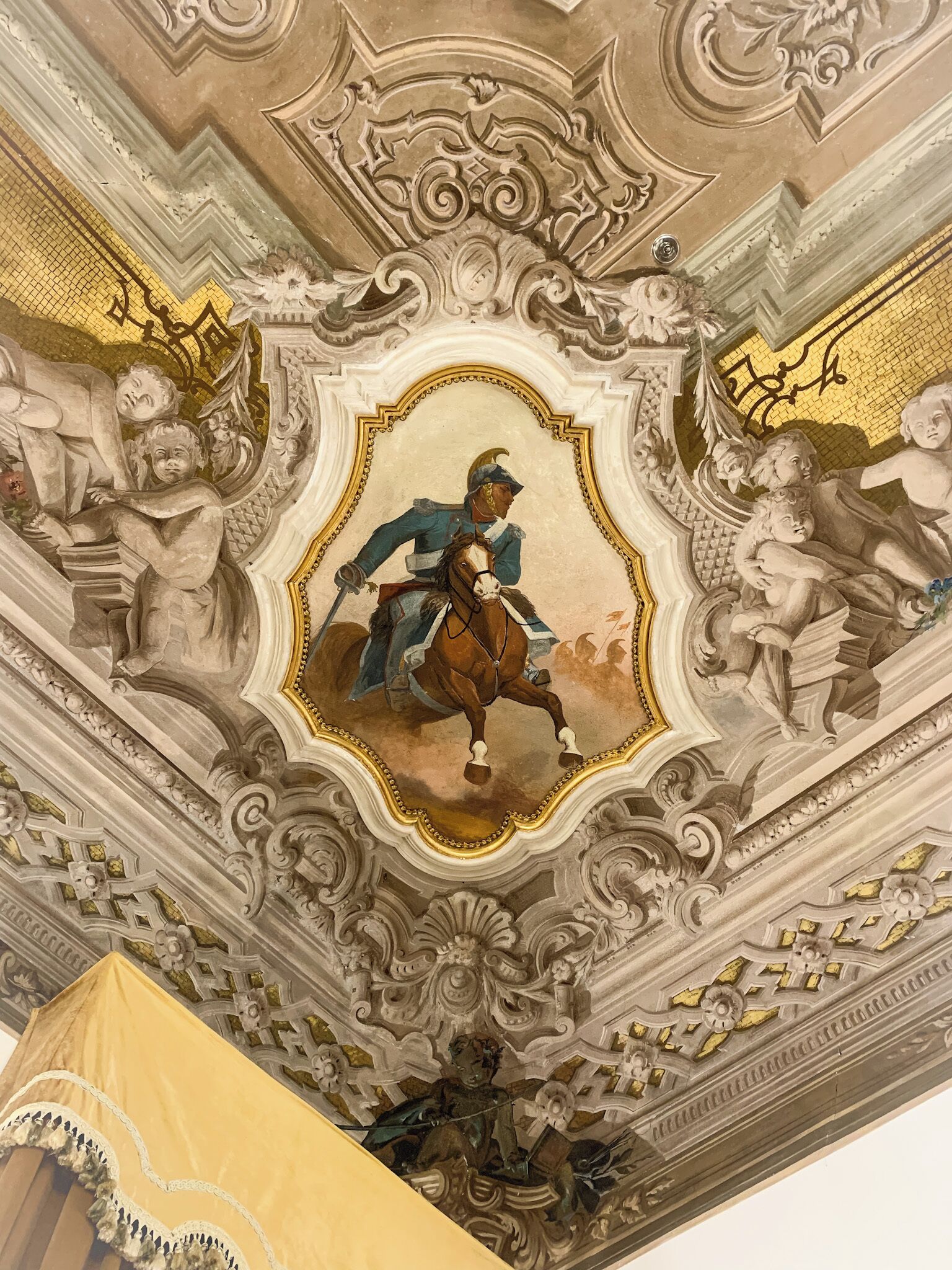 Techo detallado con un panel central ornamentado que muestra una figura a caballo, rodeado de elementos arquitectónicos y esculturas clásicas.