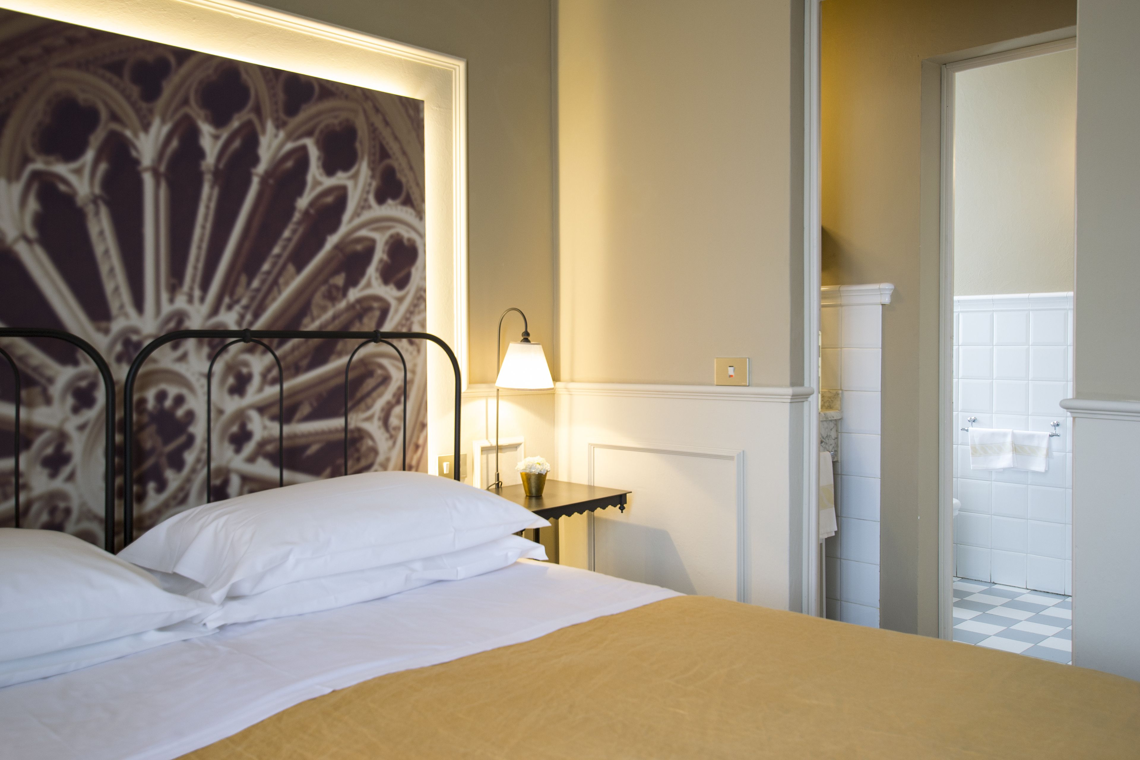 Ein Hotelzimmer mit Bett, Nachttisch mit Lampe, sichtbarem Badezimmer durch eine geöffnete Tür und einer dekorativen Wandverkleidung.