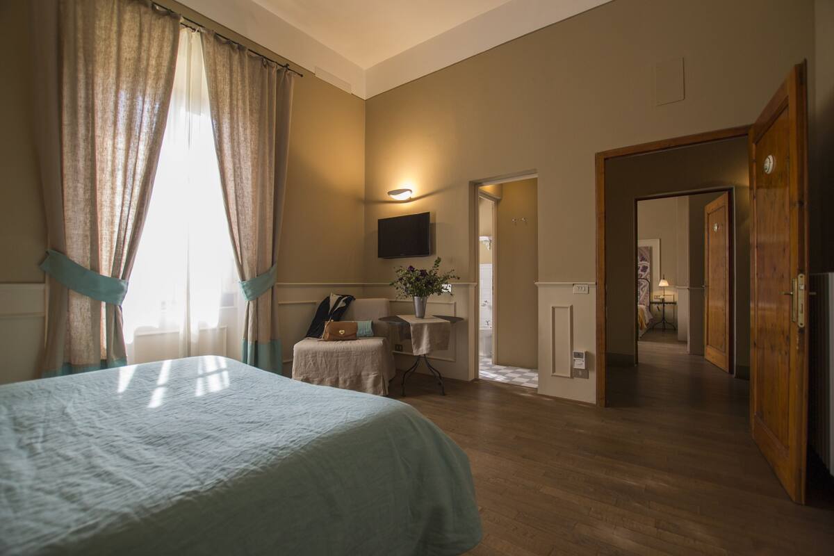 Habitación de hotel con cama, una mesita con una planta, televisor montado en la pared y una puerta de madera que da a otra habitación.