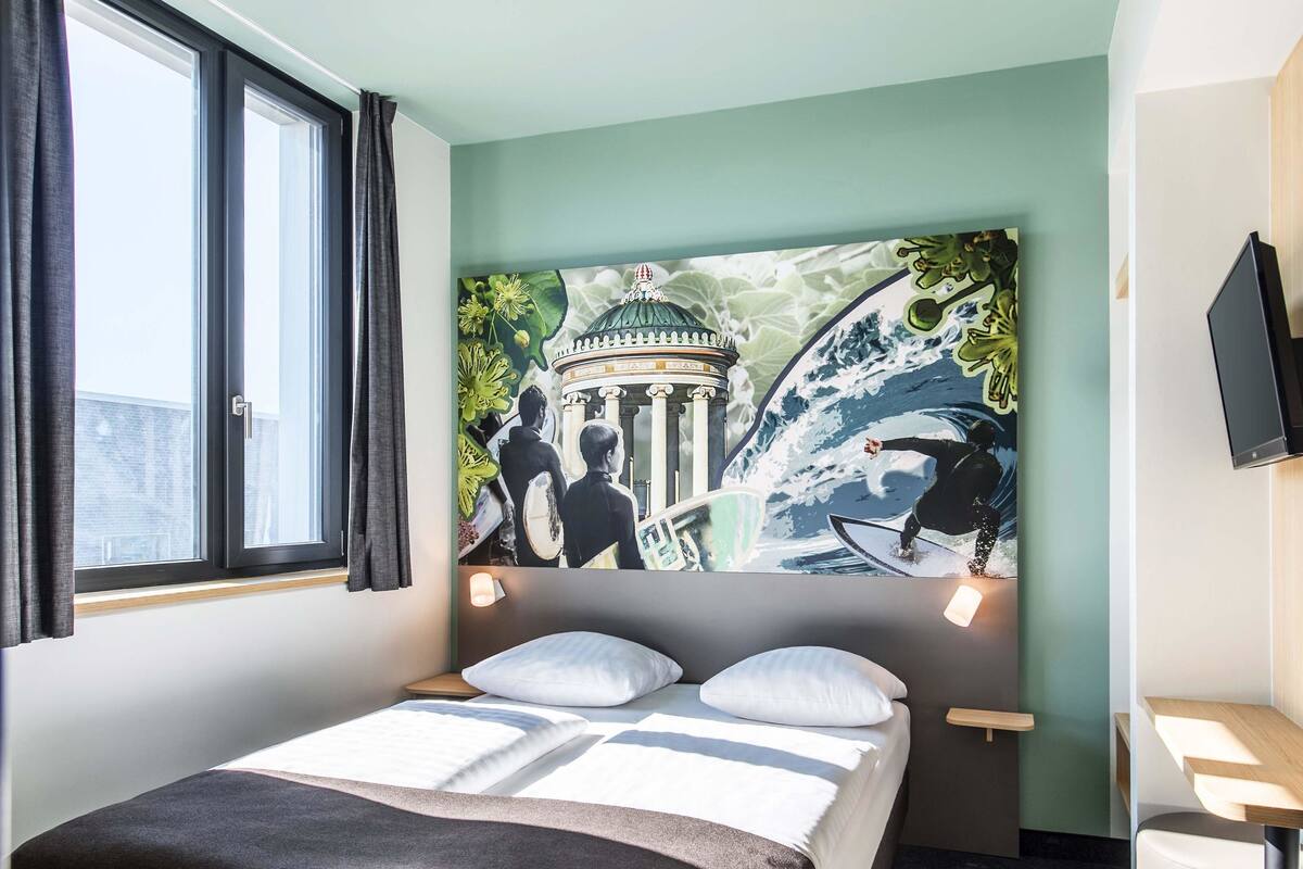 A hotel room with a bed, a window with curtains, a wall-mounted TV, and a large mural depicting a historical building and a surfer.