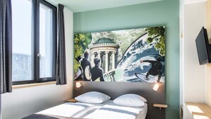 Habitación de hotel con cama, ventana con cortinas, televisor de pared y un mural grande que representa un edificio histórico y un surfista.