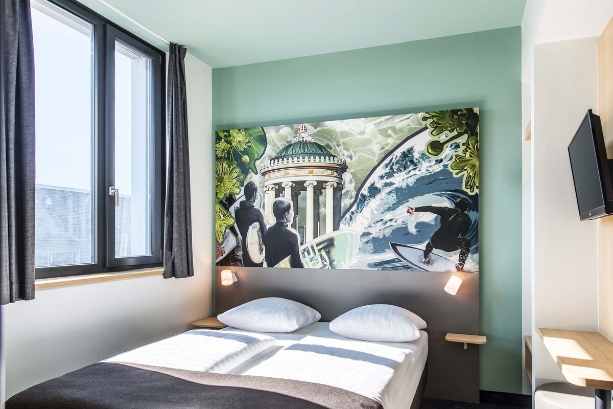 A hotel room with a bed, a window with curtains, a wall-mounted TV, and a large mural depicting a historical building and a surfer.