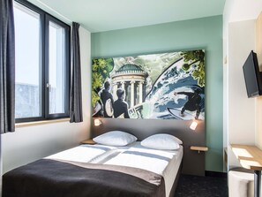 A hotel room with a bed, a window with curtains, a wall-mounted TV, and a large mural depicting a historical building and a surfer.