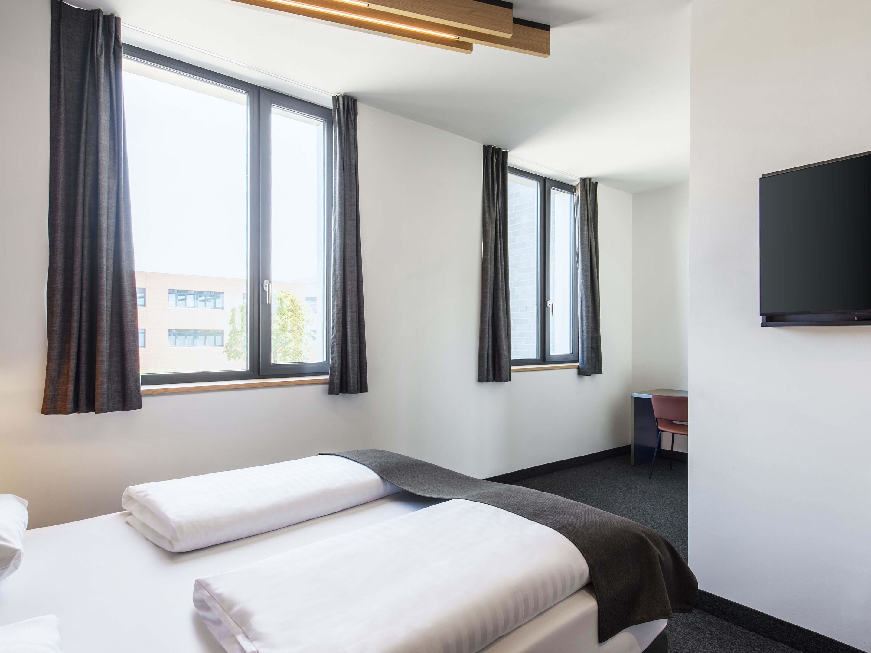 A hotel room with two beds, large windows with curtains, a flat-screen TV, and a desk.