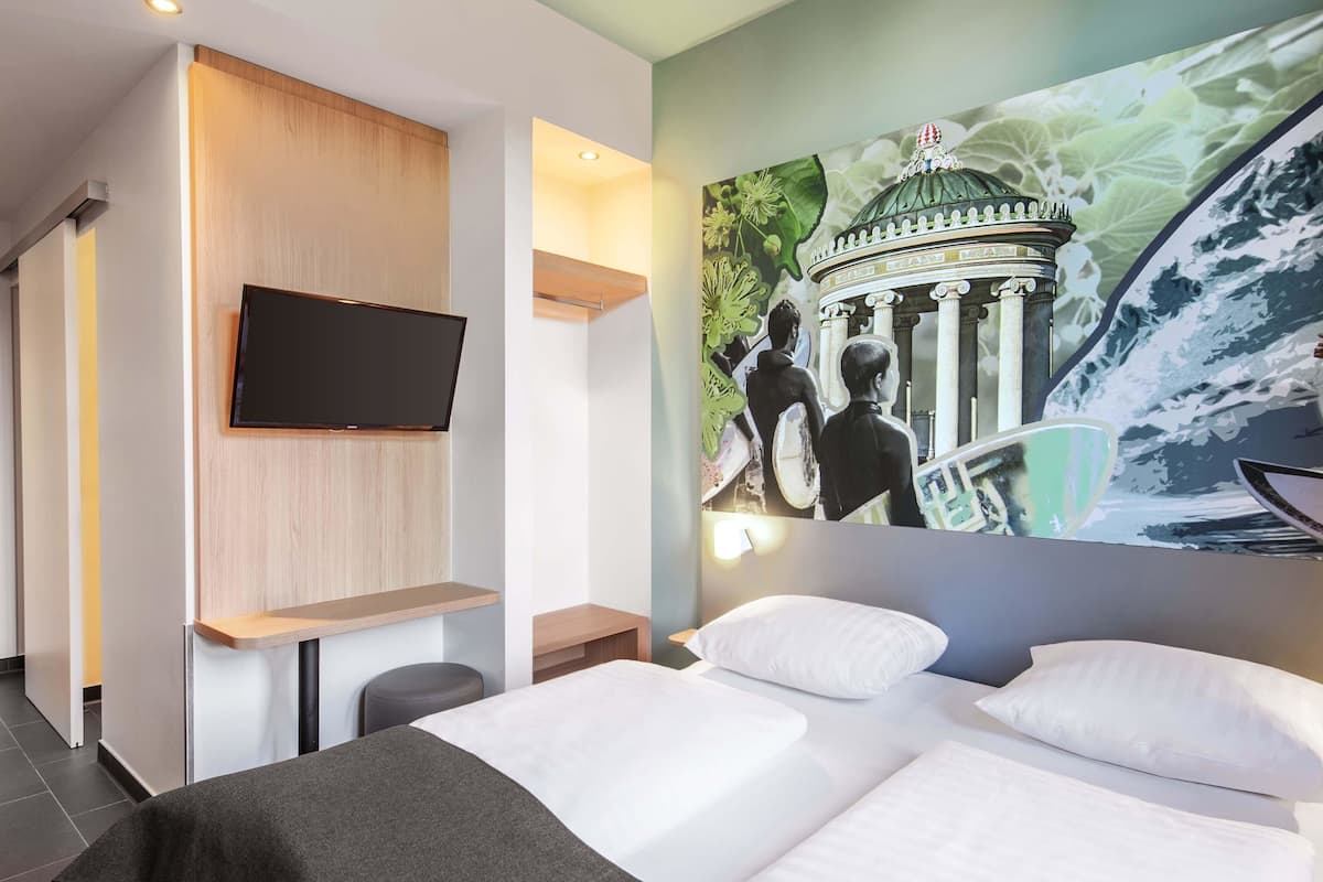A modern hotel room with a bed, a flat-screen TV mounted on the wall, and a mural depicting a historical building and two people with surfboards.