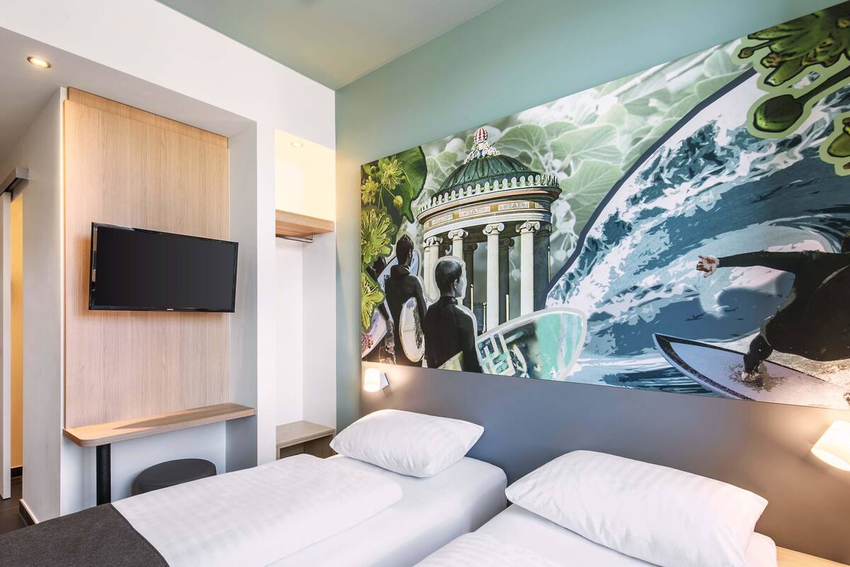 A hotel room with two beds, a flat-screen TV mounted on the wall, and a large mural depicting a surfer and a building.
